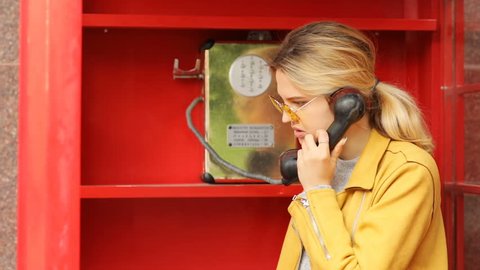 Young stylsh female person talking on phone in red call box yellow sunglasses leather jacket spring casual fashion calm serious face emotions proffesional woman calling in telephone booth film shoot