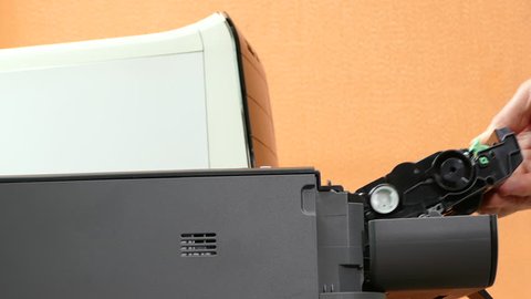 Closeup shot of a man’s hand swapping the toner cartridge in a modern compact laser printer, alongside a desktop PC.