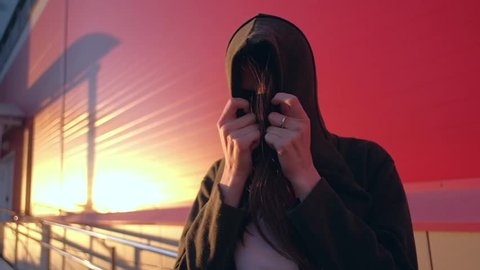 Slowmotion - mystical female take off a hood on her head looking in to camera with a strict look in red background. Urban lifestyle 库存视频