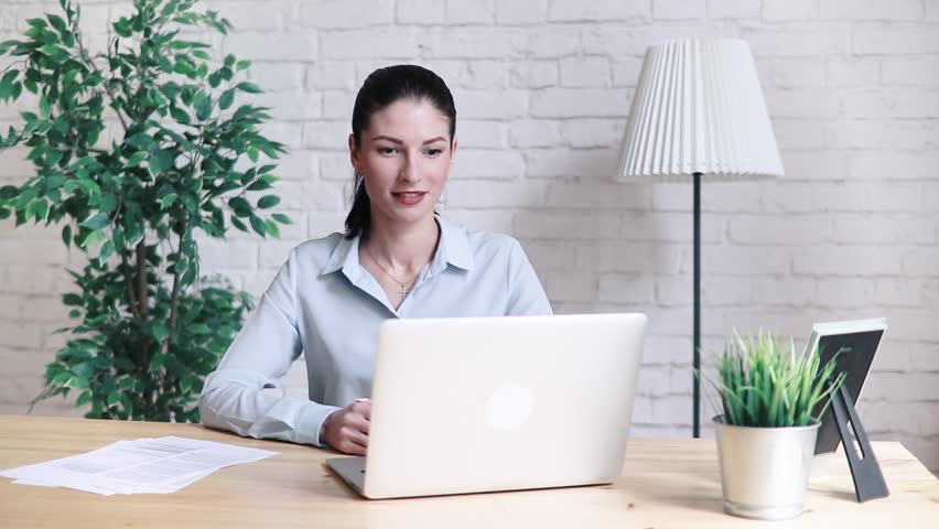 Smiling hipster girl talking and waving and Hello during a video call with a laptop sitting at the table at work. Cheerful business woman having a conference on the Internet via laptop. Royalty-Free Stock Footage #1012283036