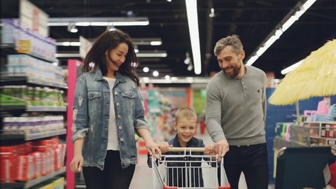 Slow motion of happy family father, mother and child running through supermarket with shopping cart, smiling and laughing. Having fun in shop and people concept.