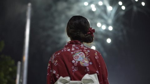 Shot from behind Asian girl in kimono waving a hand fan with fireworks in the background at a Japanese festival. Camera pans up and focus goes from girl to fireworks in 4K