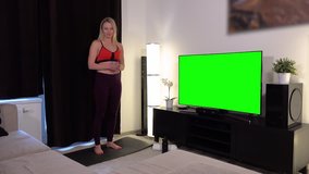 A young beautiful woman does a fitness dance in front of a TV with a green screen in an apartment and smiles at the camera