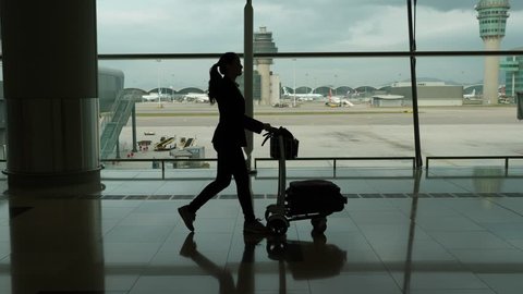Confident woman walk with carry-on luggage on small cart at international airport hall, slow motion tracking shot. Dark silhouette of professional lady wearing casual suit, arrived at HKG, going out