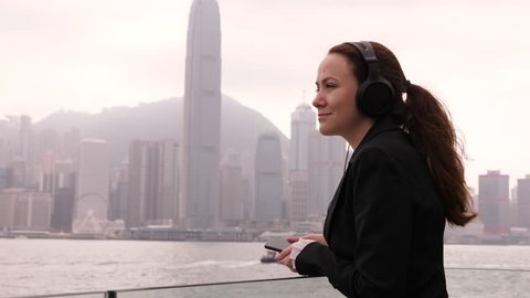 Pensive woman listen music on headphones, admire Victoria Harbour panorama, foggy Hong Kong Island buildings seen on background. Business lady have rest at famous promenade with scenic city view