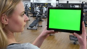 A young beautiful woman looks at a tablet with a green screen in a gym - closeup from behind