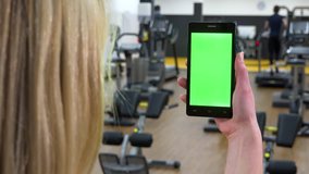 A blonde woman looks at a smartphone with a green screen in a gym - closeup from behind