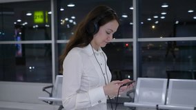 Woman watch live streaming while walking at airport sterile area, tracking shot. Lady hold smartphone in hands and listen sound through large earphones. Half length portrait shot in motion
