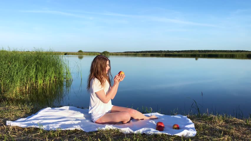 Beautiful girl in white sits by the lake and tosses an apple.