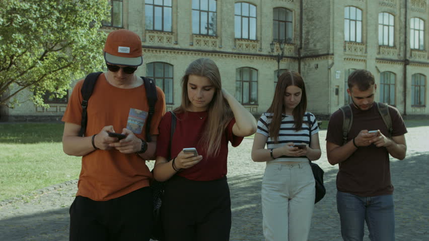Relaxed college friends busy with smart phones walking in university campus after class. High school classmates networking with cellphones while walking in campus after lecture. Stabilized shot.