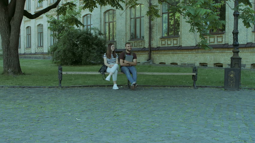 Joyful university students meeting in campus to study. Couple of friends sitting on the bench while their classmates join them in university campus. Steadicam stabilized shot.