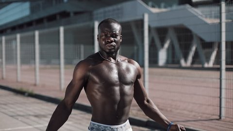 Athletic African American man with a bare torso posing shows his muscles outdoors. Black man posing near the stadium, shows his muscles. Bodybuilding, posing, health, fitness, beauty.