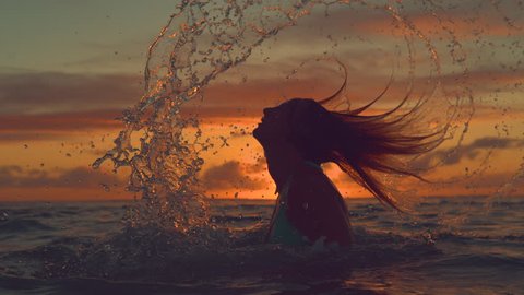 SLOW MOTION, CLOSE UP, SILHOUETTE: Playful girl splashes refreshing sea water with her long hair on a spectacular summer evening near tropical island. Amazing shot of woman flipping her hair back.