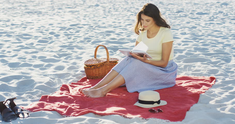 Young attractive Caucasian woman flipping pages of the book while sitting on the seacoast and reading on a nice summer day. Outside.