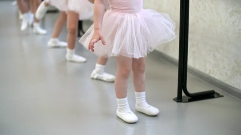 Tilt up shot of group of little girls in ballet clothes standing at ballet barre and stretching legs for split, their female teacher helping them