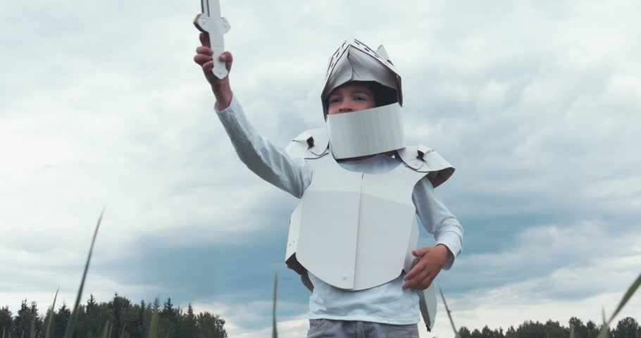 Kid boy wearing cardboard medieval knight armor costume draws a sword from a scabbard against blue sky. 4K UHD 60 FPS SLO MO