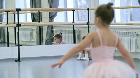 Sad little girl sitting on the floor in dance studio and looking at other girls having ballet lesson