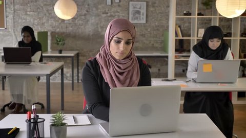 Three young muslim womans in hijab sitting and working in modern office, muslim beautiful woman looking in camera and smiling