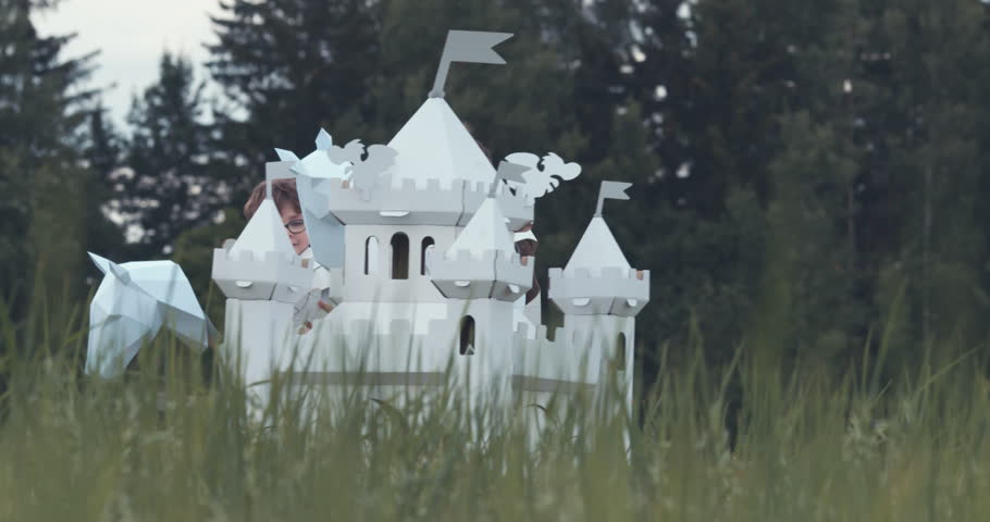 Brothers boys wearing cardboard medieval knight armor costumes riding stick horses through a grass field in summer. 4K UHD 60 FPS SLO MO