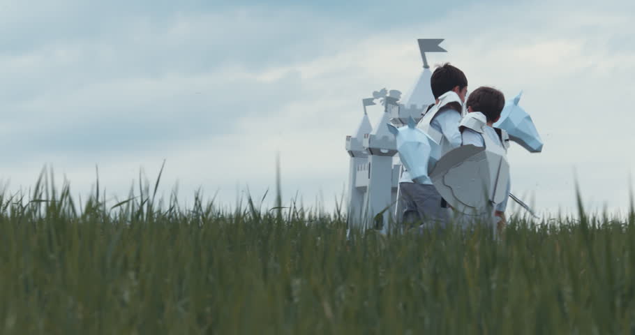 Brothers boys wearing cardboard medieval knight armor costumes riding stick horses through a grass field in summer. 4K UHD 60 FPS SLO MO