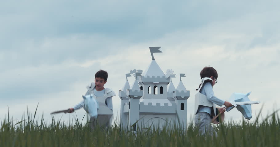 Brothers boys wearing cardboard medieval knight armor costumes riding stick horses through a grass field in summer. 4K UHD 60 FPS SLO MO