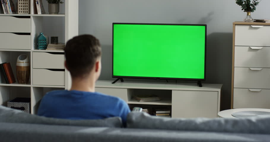 Back view on the young man sitting on the couch in the living room, watching TV with green screen and changing channels with a remote control in his hand. Chroma key. TV in focus. Indoor Royalty-Free Stock Footage #1012385369