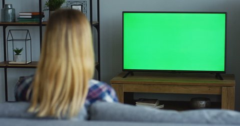 Rear of the blonde woman in the plaid blue shirt sitting on the sofa in the living room and watching TV with green screen, then changing channels with a remote control. Chroma key. Indoors