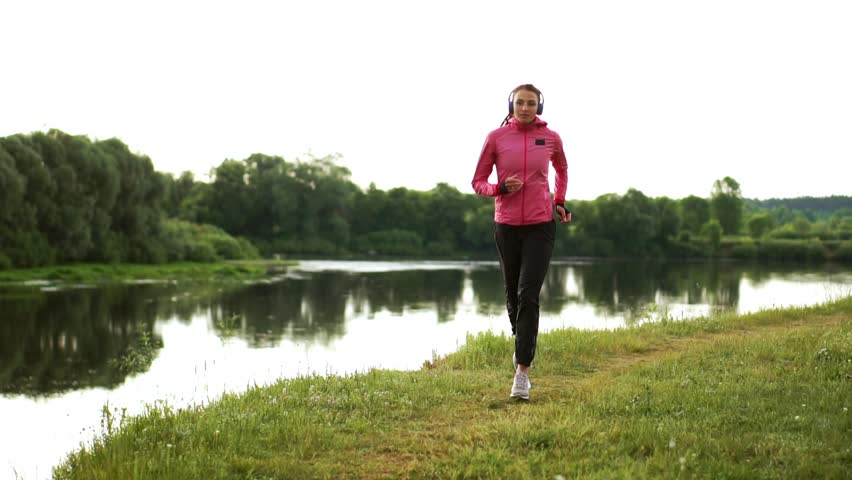 Brunette with long hair in headphones runs along the river in the Park in the morning at sunrise in the summer in a pink jacket and black pants