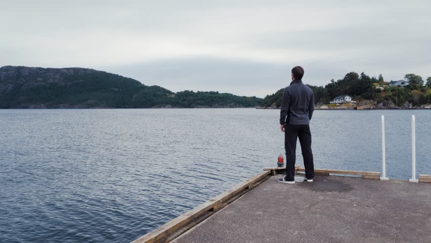 Male tourist meditatively peering at beautiful landscape. Amazing pond, lake, sea, calm water surface. High mountains covered green forest, clear blue sky. Small town, settlement in Norway. Zoom in.