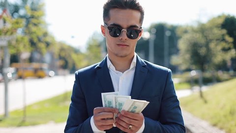 Young happy businesman in sunglasses and a suit counting money and walking in the street. He selebrating his successful win with a lot of dollars
