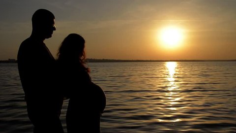 pregnant woman with her husband standing near the river at sunset