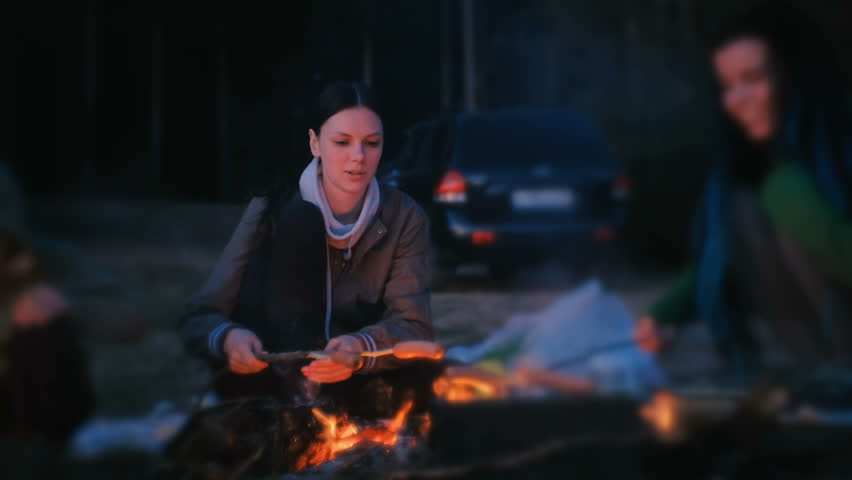 Two women fry sausages on a fire. Family camping at night.