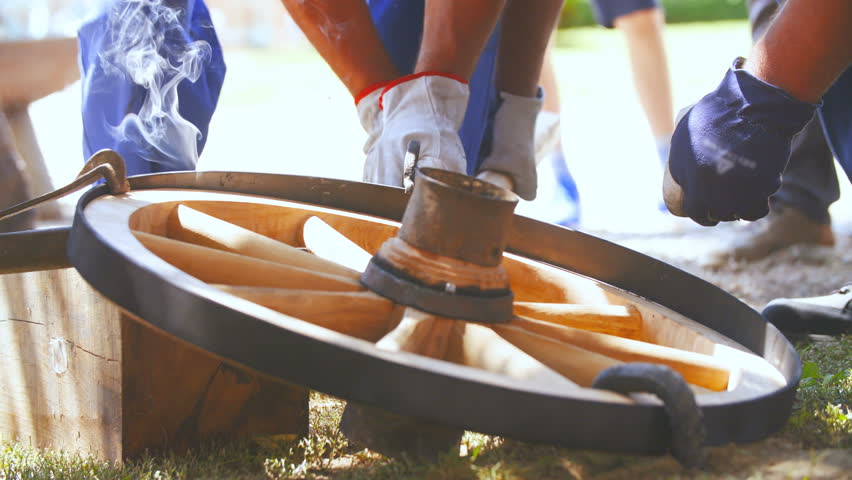 Blacksmith protects wooden carriage wheel with hot iron close-up 4K. Low angle long shot of two persons hands in focus putting together iron ring with wooden wheel.