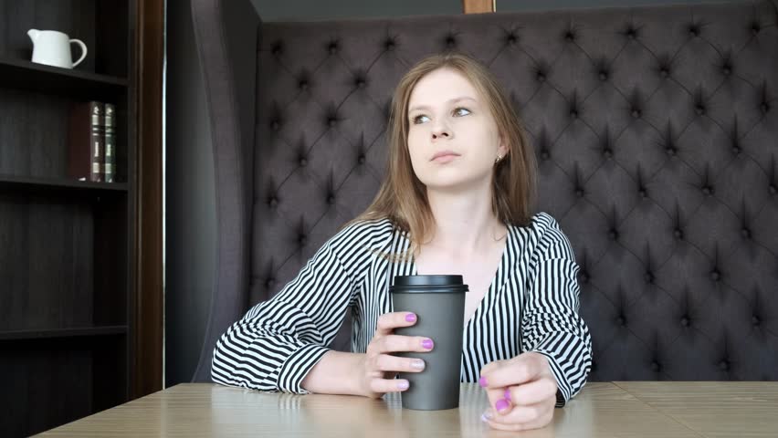 A young beautiful girl in a stylish cafe drinks coffee in the morning before work. Sits opposite the window. He drinks coffee from a cardboard black glass. Close-up