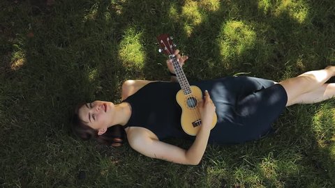 Young woman resting, she play on ukulele, little guitar, lying on grass
