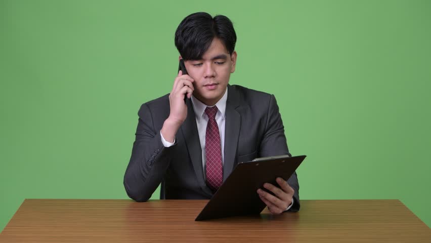 Young handsome Asian businessman working with clipboard against green background