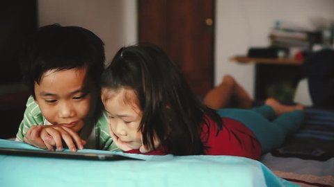Pan video of asian children looking at digital tablet. Focus at chinese boy, younger sister lying prone on bed, near by. Conceptual about using E-learning technologies. Cinematic tone.