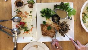 Woman hand holding knife and fork cutting grilled beef steak in white dish with food set and another is finish on the wooden table at restaurant. Top view. video footage.