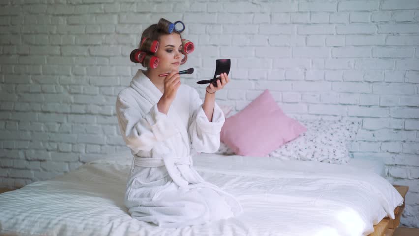 Makeup young woman putting lipstick wearing hair rollers getting ready for going out.