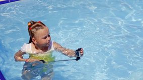 baby girl standing in the pool in the water and sets up an action camera on a selfie stick to record her personal video blog