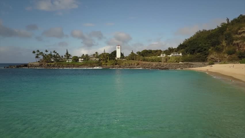 Waimea Bay Oahu hawaii. Drone view of white sandy beach, turquoise water, waves and church. Sunny weather and people swimming. Cars drive by on road. 