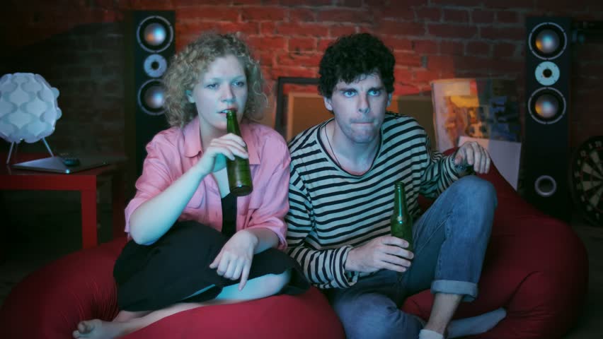 Young married couple enthusiastically watching the live broadcast of the football championship, eating chips, drinking beer and shouting enthusiastically - hurray, goal
