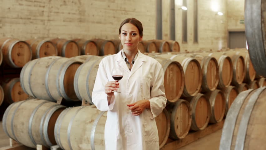 Young cheerful woman wearing coat holding glass of wine in large cellar on winery factory

