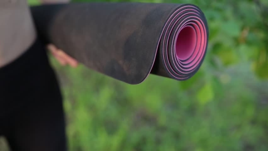 Young woman rolling her yoga exercising mat after a yoga practice on green grass in the park at sunset, close up