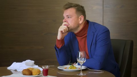 Tired businessman sitting at table in restaurant during dinner. Young man does not want to eat unappetizing dishes in cafe during business lunch