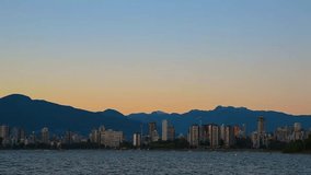 Video panorama from the sea to a modern city with mountains and tall buildings, a huge blue sky with a glow of sunset, yachts and boats on the water, driving cars on a summer evening