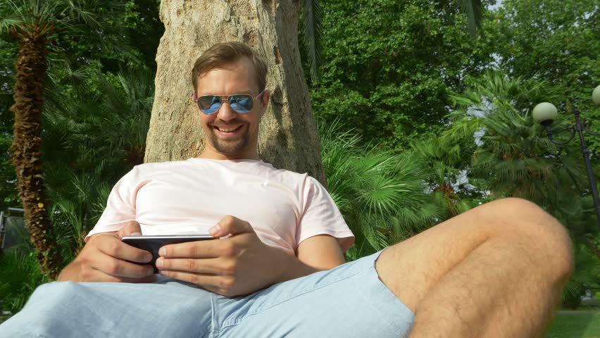 Young tourist sitting under a palm tree with a mobile phone. Positive emotions. Time relax. 4k, slow motion