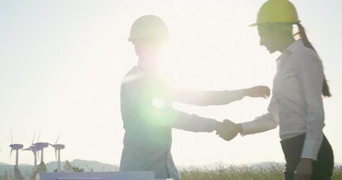 Two engineers talk about the project for the construction of a renewable energy wind farm; the two shake hands with the finished work in a wind farm at sunset.