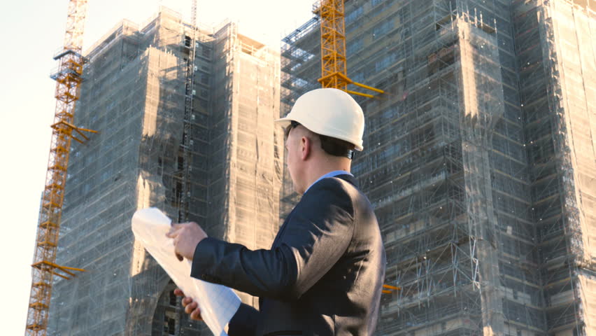 Portrait of a successful young handsome engineer, architect, builder, businessman, wearing a white helmet, in a suit, holding a project in his hand, a skyscraper background and a construction site. Royalty-Free Stock Footage #1012510889