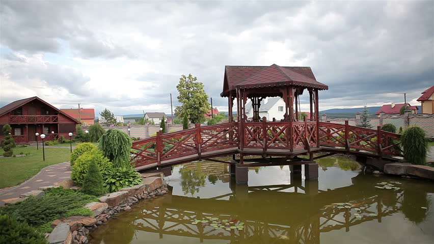 A wooden bridge with a little house over the lake. Colorful fish swim in the water. Thunderclouds are floating in the sky.